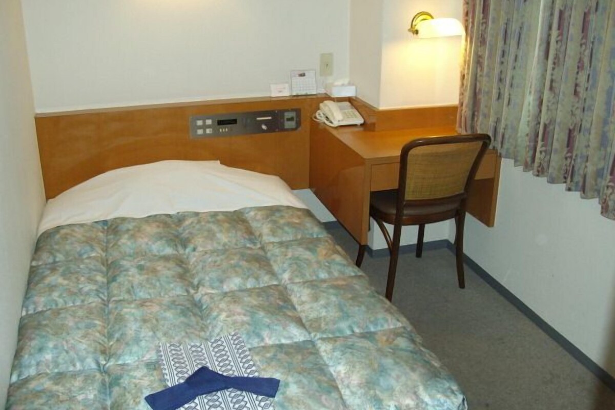 A hotel room with a bed, a desk, a chair, a telephone, and a wall-mounted light fixture.