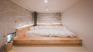A minimalist bedroom with a wooden platform bed, a concrete wall, and a small window. - Yoshida Mura Inn (Shimotsuke)
