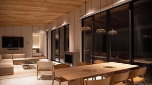 A modern living room with a wooden ceiling, a large window, a sofa set, a dining table with chairs, and a flat-screen TV mounted on the wall. - Soothe Forest (Awaji)