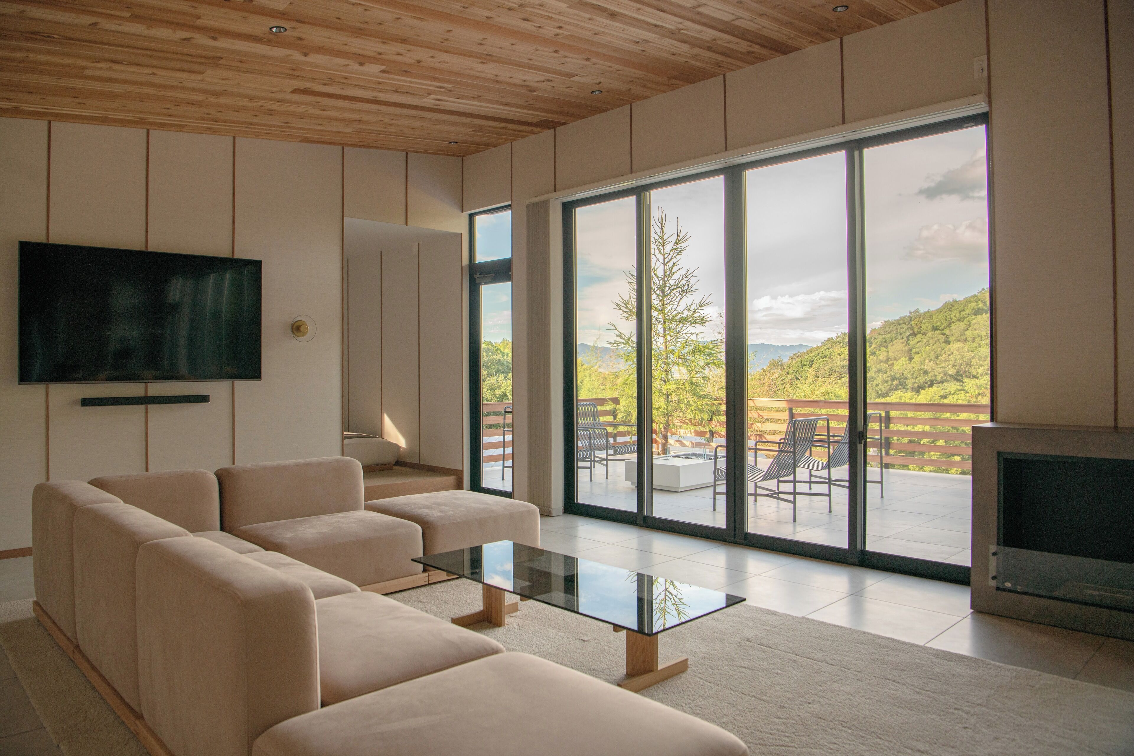 A modern living room with a large TV, beige sofas, a glass coffee table, and a balcony with outdoor furniture.