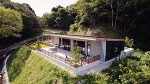 A modern house with a flat roof, large glass windows, and a patio area with outdoor furniture. - Soothe Forest (Awaji)