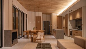 A modern living room with a glass coffee table, wooden furniture, and a kitchen area with a countertop and appliances. - Soothe Forest (Awaji)