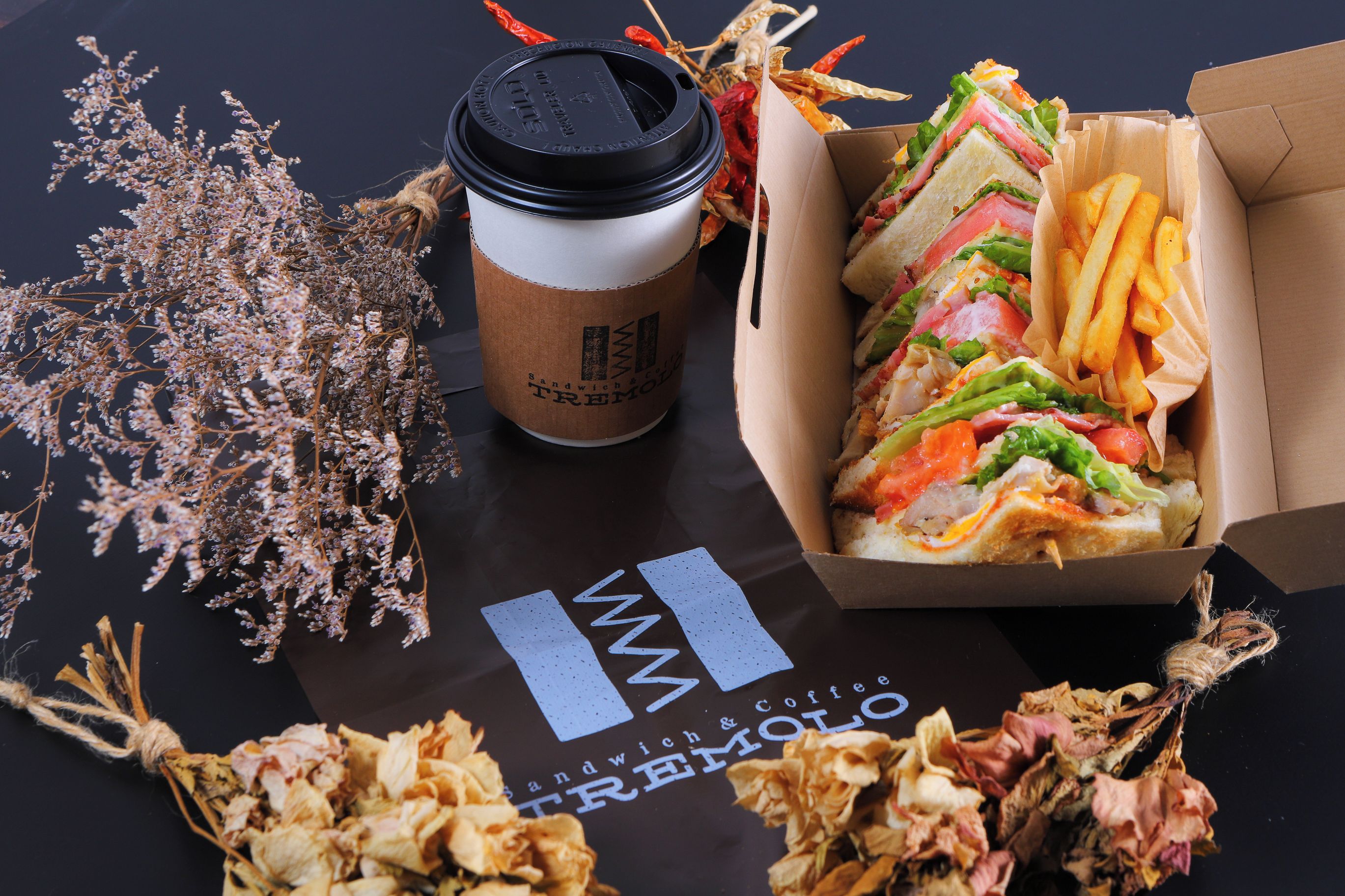 A sandwich, fries, a coffee cup, and dried flowers on a table with a logo.