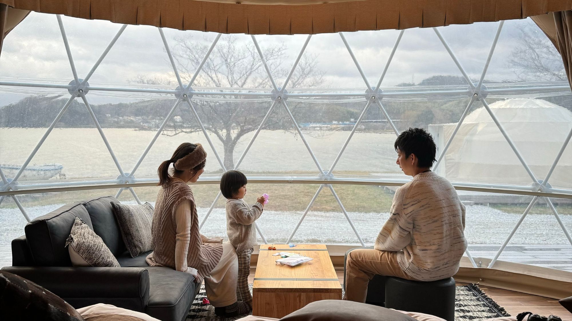 A room with a large window, a black leather sofa, a wooden table, and a view of a body of water and hills.