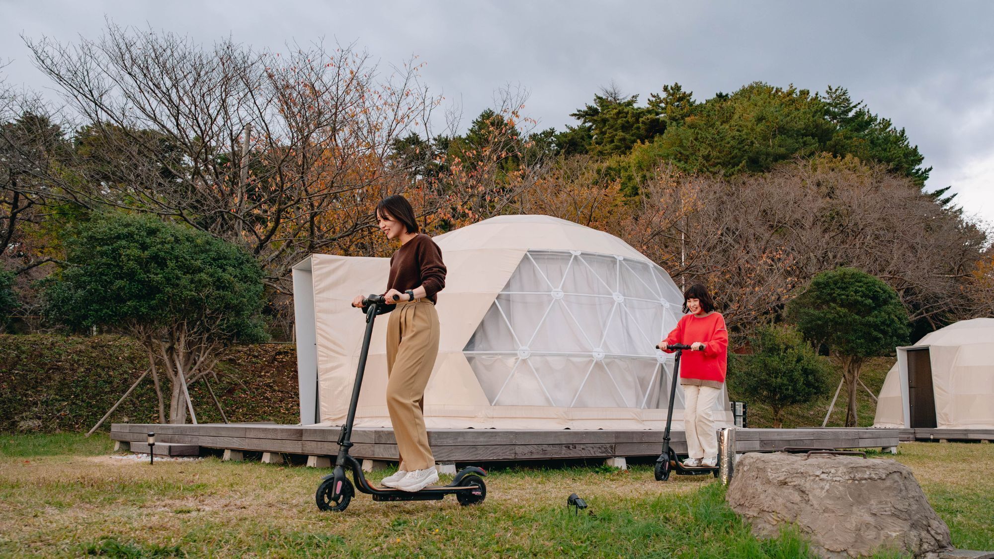 Two people on electric scooters in front of a large, dome-shaped tent-like structure.