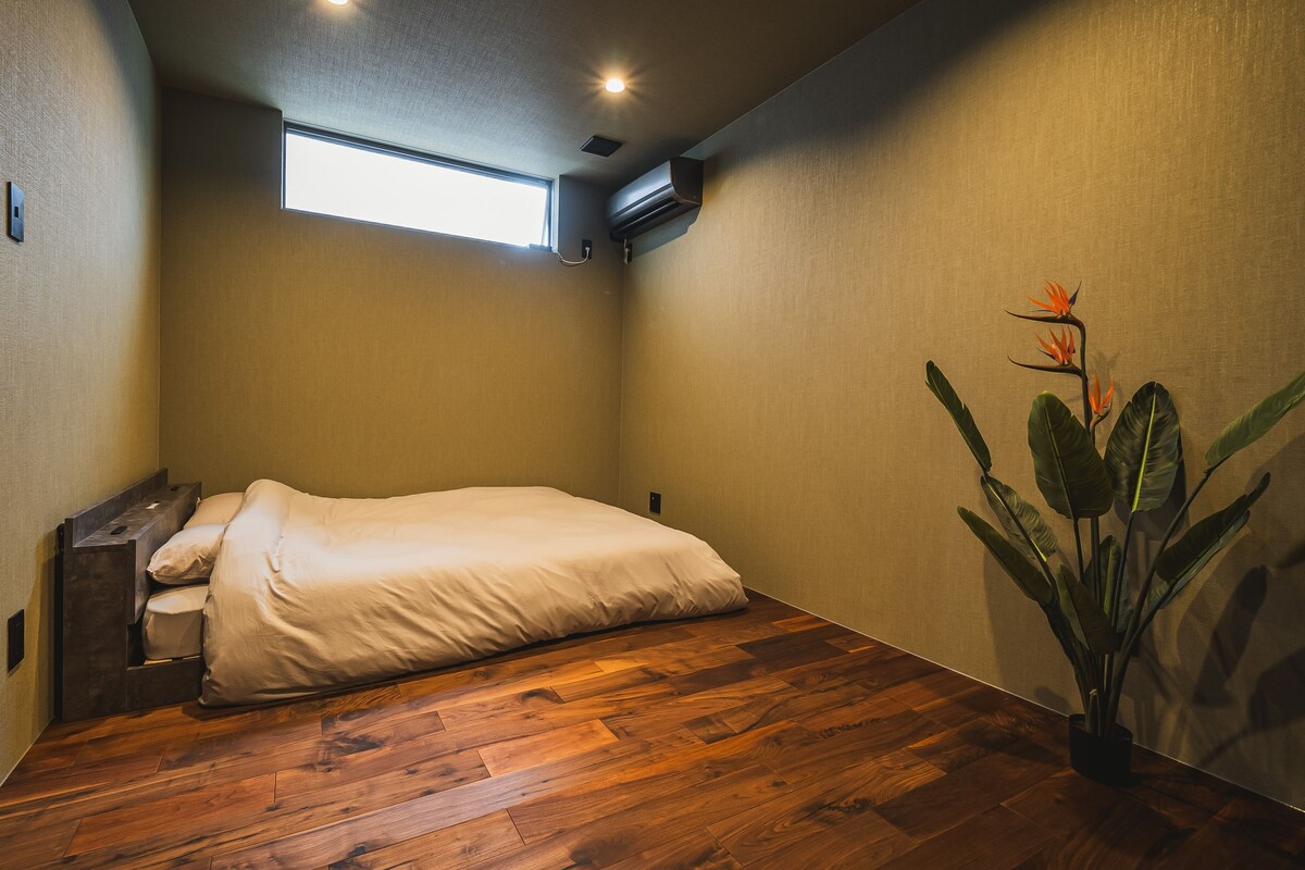 A small, cozy bedroom with a wooden floor, a bed, a nightstand, a window, and a potted plant.