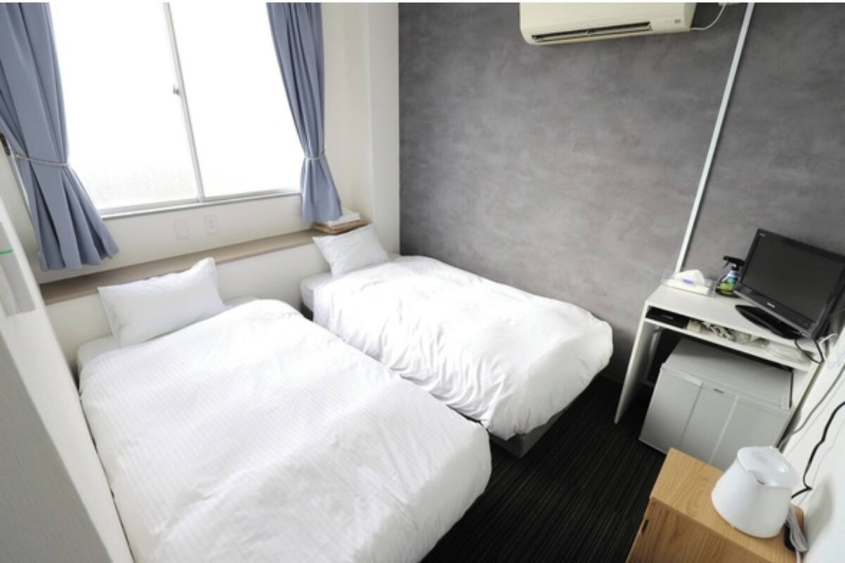 A hotel room with two beds, a desk with a computer, a small fridge, and a window with curtains.