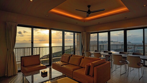 A spacious living room with a large sofa, a coffee table, and a dining area with chairs. The room features large windows with a view of the sea and a balcony. - United Resort Chatan Ocean Front (Chatan)
