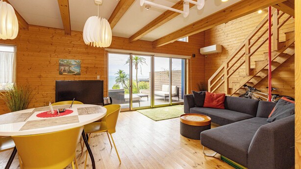 Ein modernes Wohnzimmer mit Holzvertäfelung, einem Essbereich mit gelben Stühlen, einem grauen Sofa und einer Treppe.