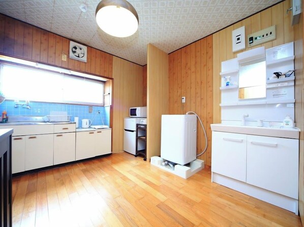 A compact kitchen with wooden paneling, a microwave, a water heater, and a window providing natural light. - Kamon Inn Kotobuki (Izumi)