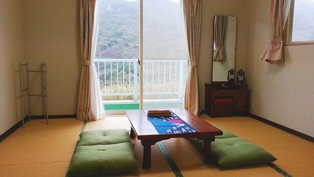 Una habitación con suelo de tatami, una mesa baja, cojines verdes, un armario de madera y un ventanal corredero que da a un balcón con vistas a un paisaje montañoso.