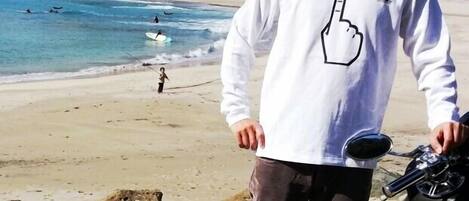 Escena de playa con una persona sosteniendo una cámara y un casco, una tabla de surf y un niño caminando por la orilla.