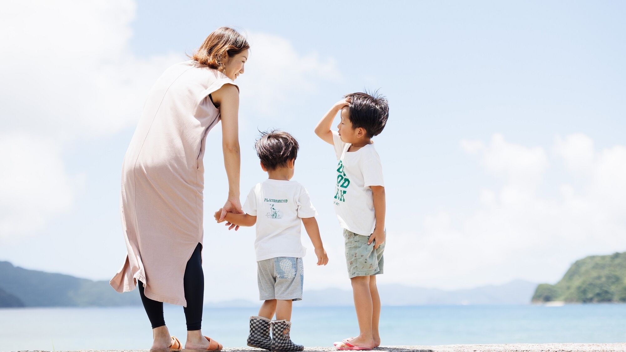 Una donna e due bambini che si tengono per mano su una spiaggia, con un cielo sereno e un mare calmo sullo sfondo.