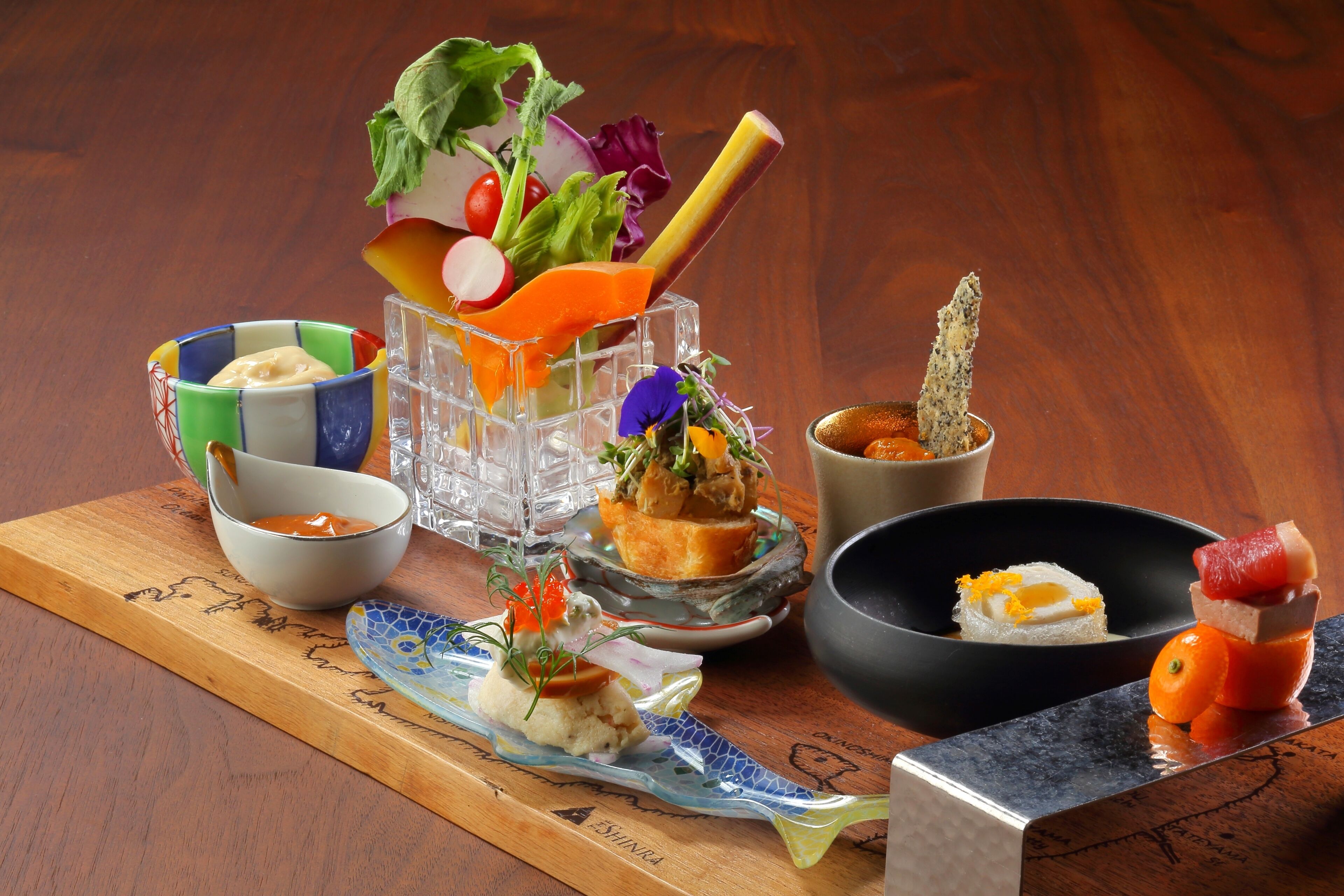 A variety of dishes on a wooden board, including a bowl of soup, a plate with a fish and vegetables, and a small dish with a sauce.
