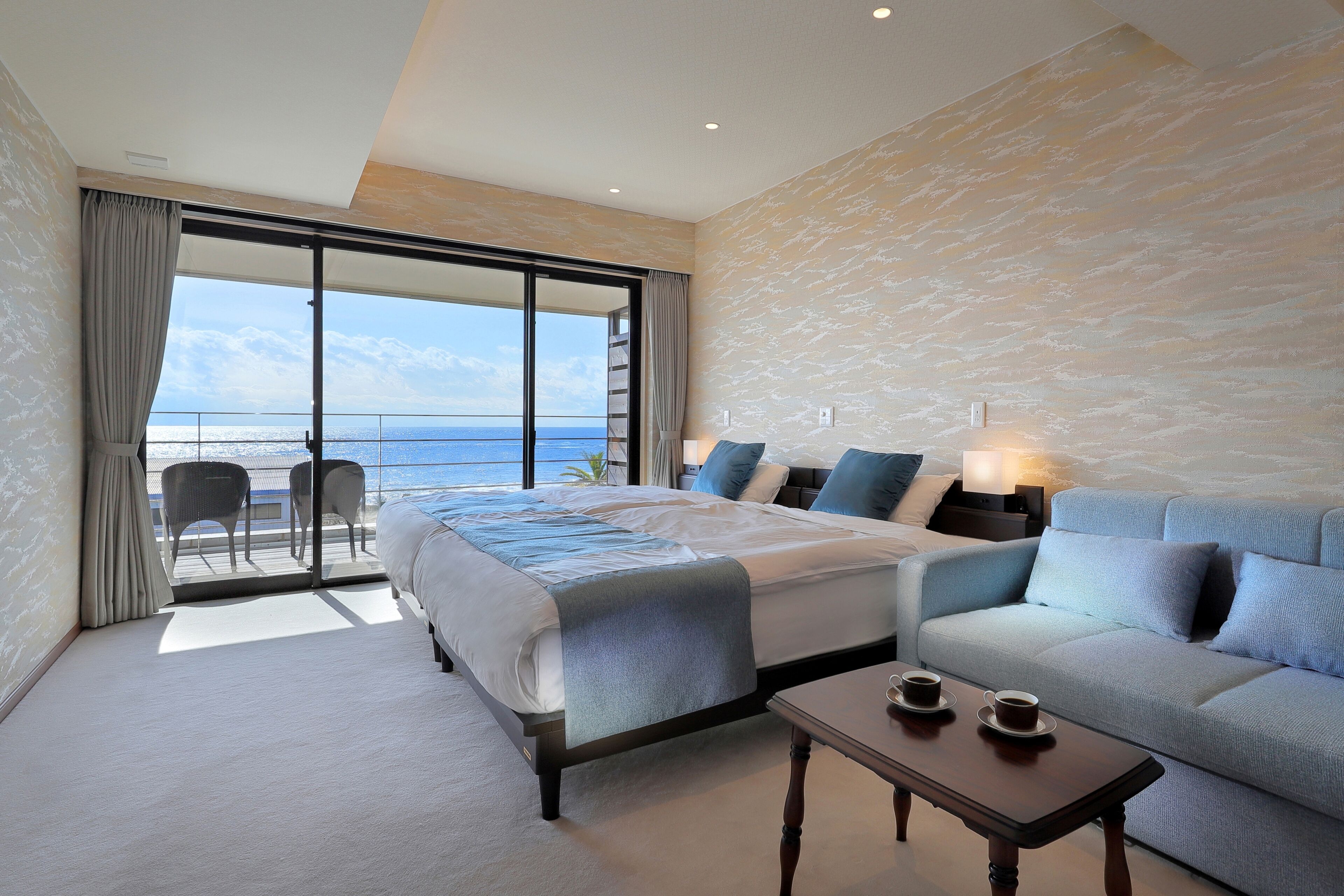 A hotel room with a bed, a sofa, a coffee table, and a balcony with a view of the sea.