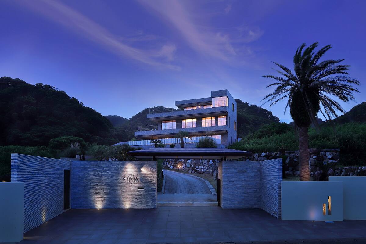 A modern two-story building with a glass facade, surrounded by lush greenery and a palm tree.