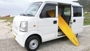 A white van parked on a concrete surface with a yellow surfboard propped against the side. - Guesthouse Ryofu (Amami)