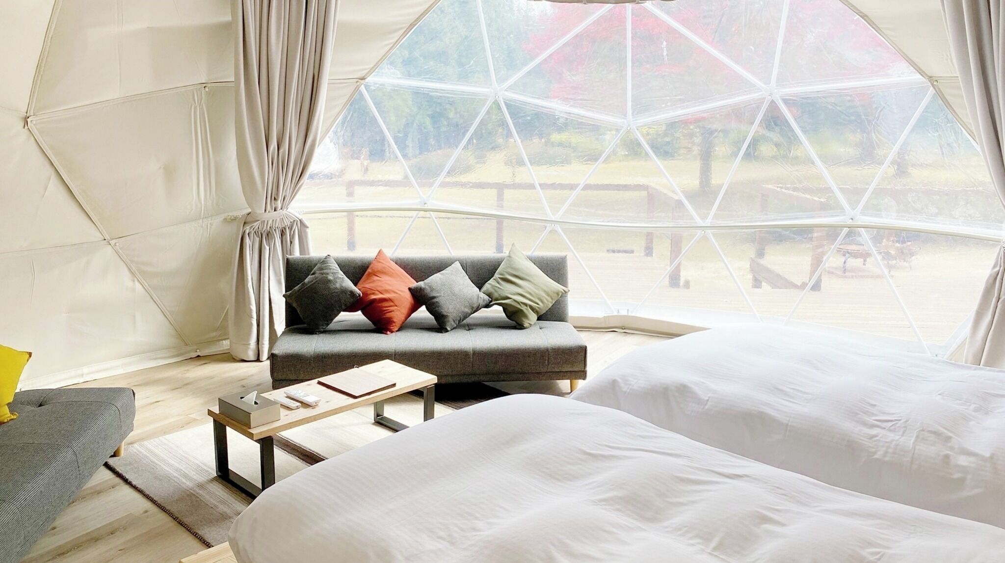 A tent-style room with a sofa and two beds, a wooden table, and a view through a large window.