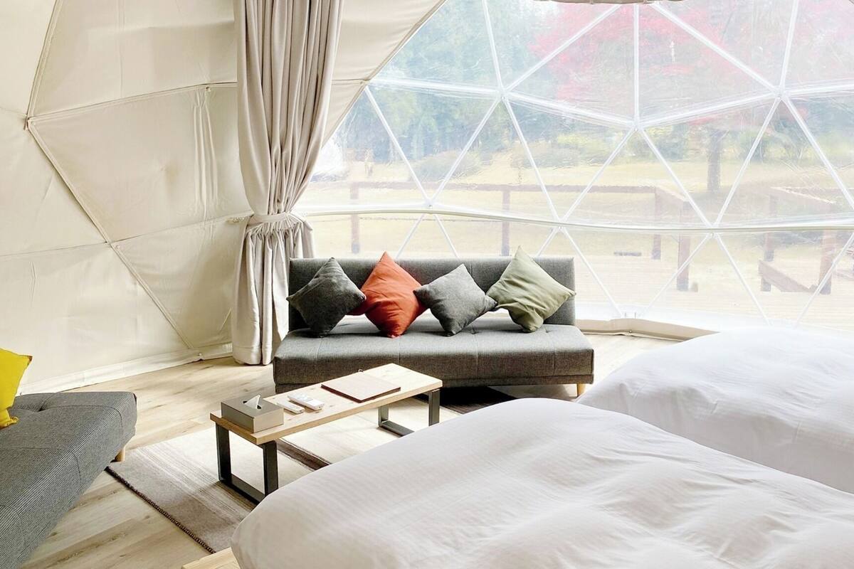 A tent-style room with a sofa and two beds, a wooden table, and a view through a large window.