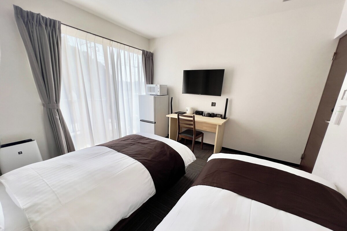 Una habitación de hotel con dos camas, un escritorio, un televisor de pantalla plana y un ventanal con cortinas.