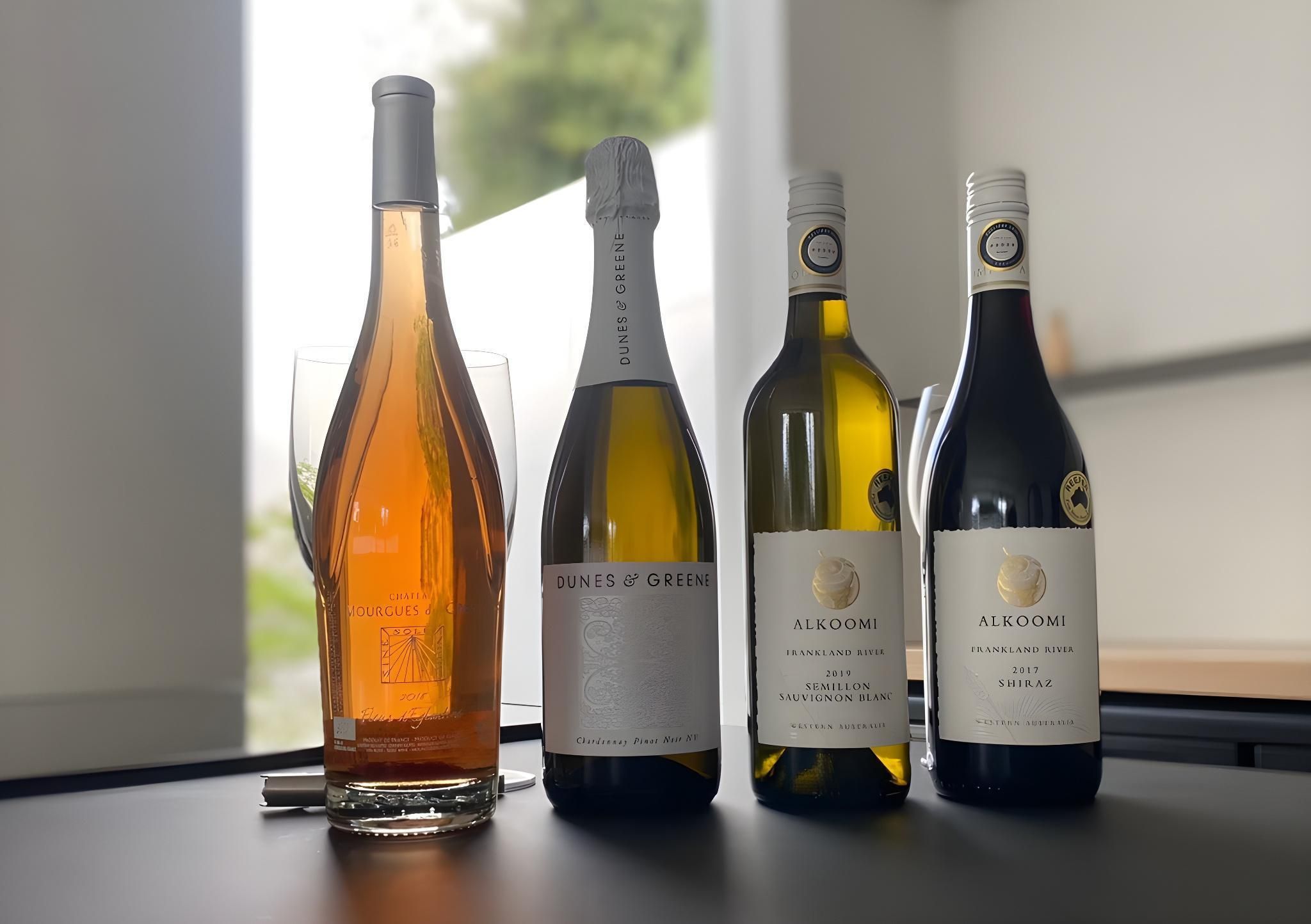 Four wine bottles and a wine glass on a table by a window.