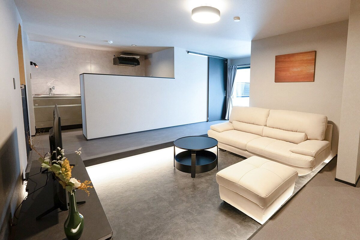 Un soggiorno moderno con un divano, un poggiapiedi e un tavolino rotondo. C'è una TV a schermo piatto su un mobile, un vaso con fiori e un quadro appeso al muro.