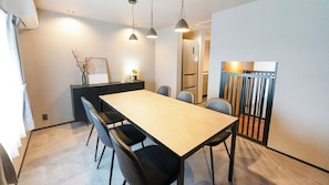 A modern dining area with a long table, black chairs, pendant lighting, and a kitchen in the background. - Izumo Dogs (Izumo)