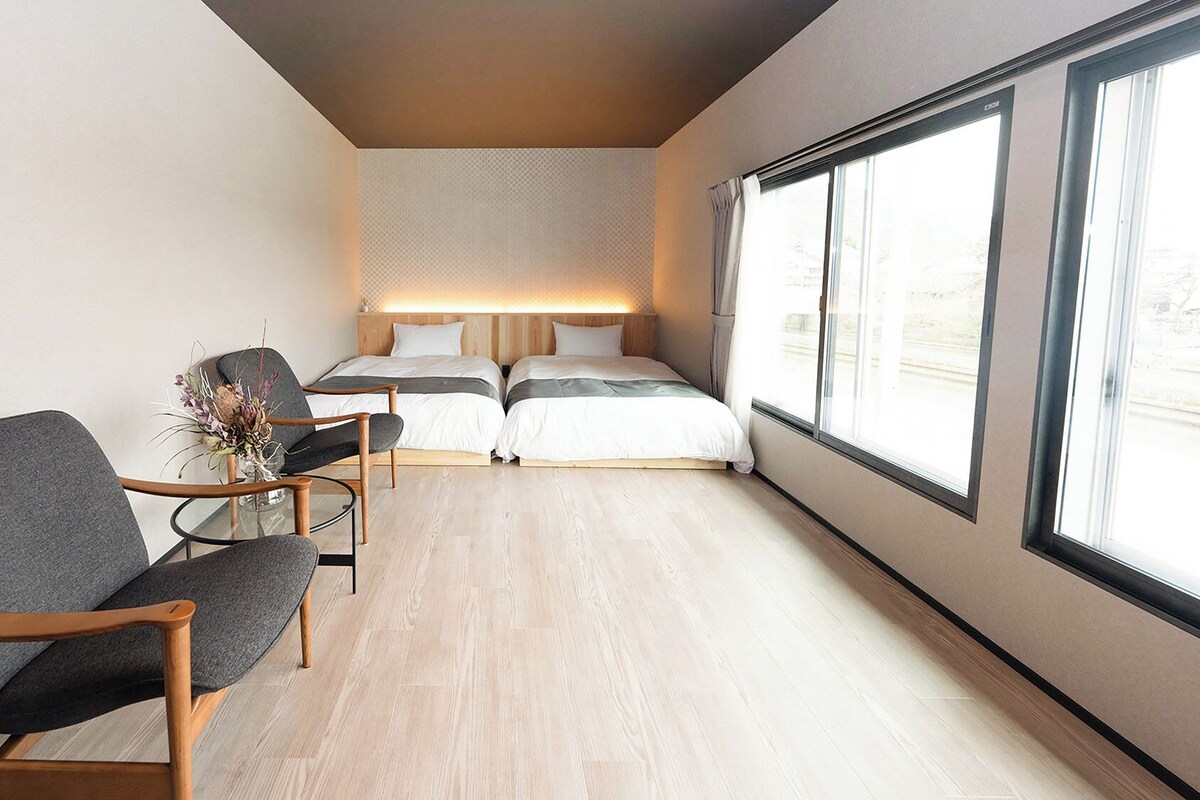 Una moderna camera d'albergo con una grande finestra, un letto con testiera, un angolo salotto con una poltrona e un tavolino, e un vaso con fiori.