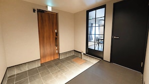 A hallway with a wooden door, a black door, and a window with a view of a room with chairs and a table. - Izumo Dogs (Izumo)