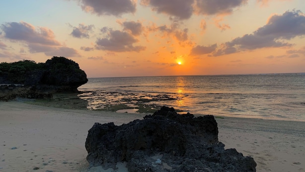 Een zonsondergang boven een strand met rotsformaties en rustig water.