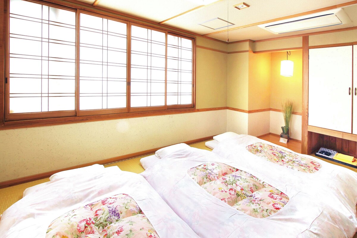 Una habitación japonesa tradicional con suelo de tatami, dos camas con ropa de cama floral, un puertas correderas y una pequeña mesa con una planta.