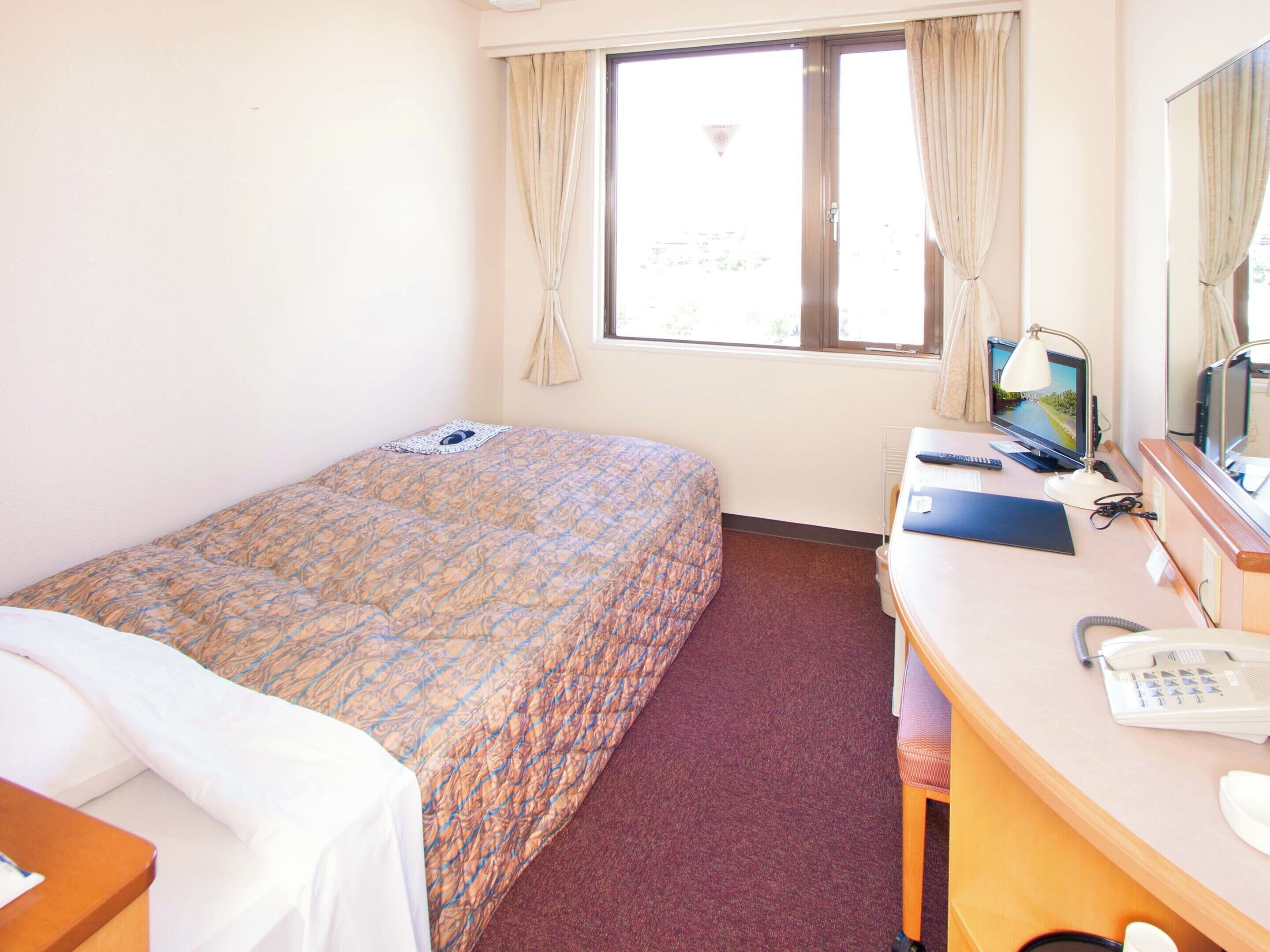 Ein Hotelzimmer mit einem Bett, einem Schreibtisch mit Computer, einem Telefon und einem Fenster mit Vorhängen.
