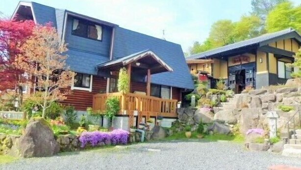 A two-story house with a sloped roof, a wooden deck, and a stone pathway leading to the entrance. - Yunomori Kusu (Kusu)