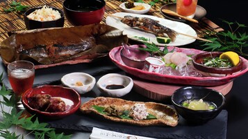 Diversos platos, incluyendo pescado a la parrilla, arroz y verduras, servidos sobre una mesa de pizarra negra con un tapete de bambú al fondo.
