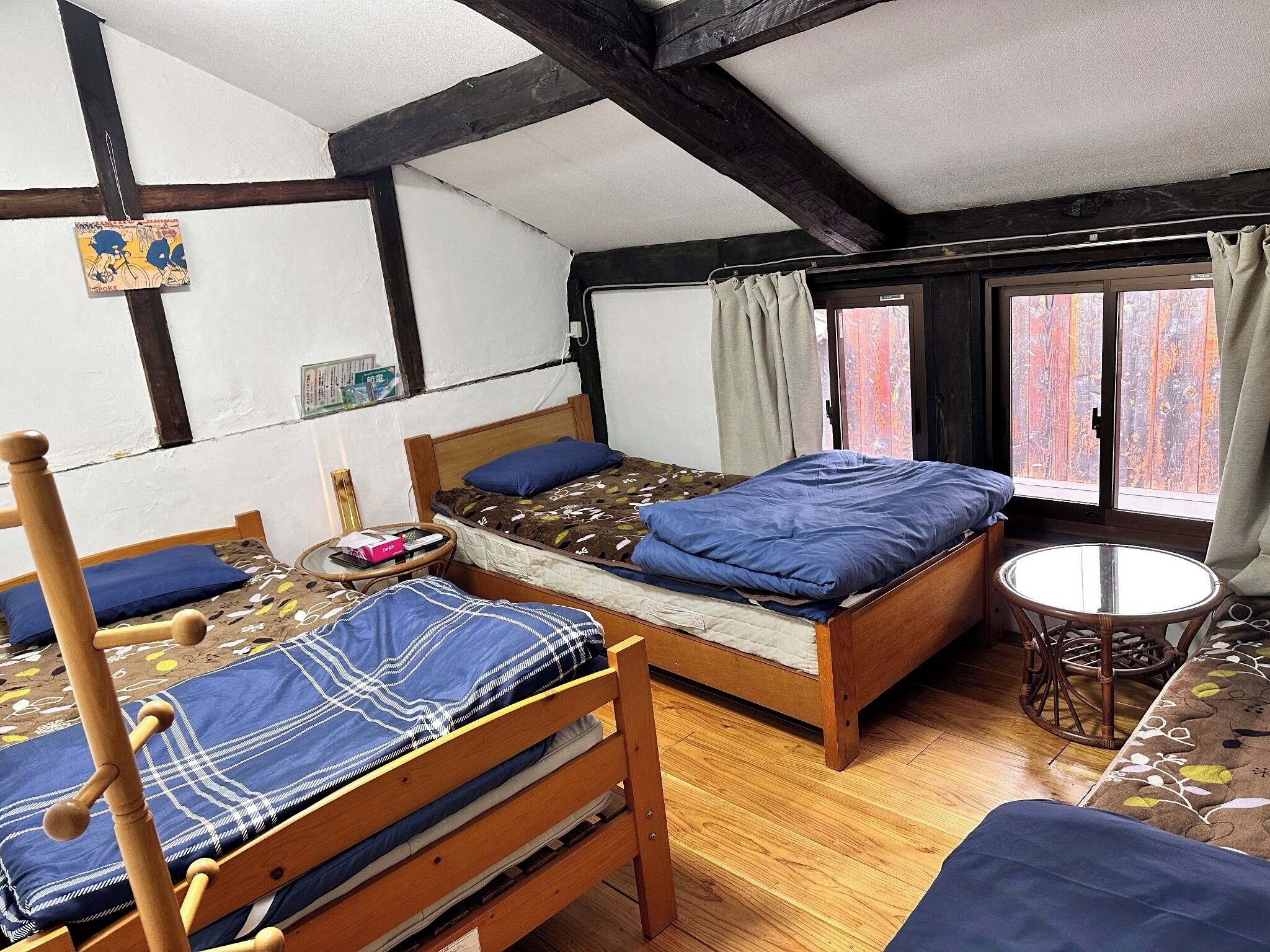 Una habitación con dos camas de madera, una mesita redonda pequeña y un ventanal con cortinas.