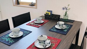 Une table à manger est dressée avec quatre services à thé, chacun comprenant une tasse, une soucoupe et une cuillère. Des chaises entourent la table, et une fenêtre se trouve au-dessus.