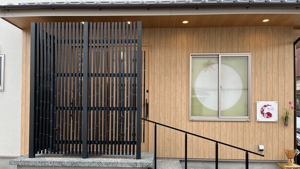 L’entrée d’un immeuble se caractérise par une façade en bois, un portail en métal noir et un escalier en béton menant à une porte en bois.