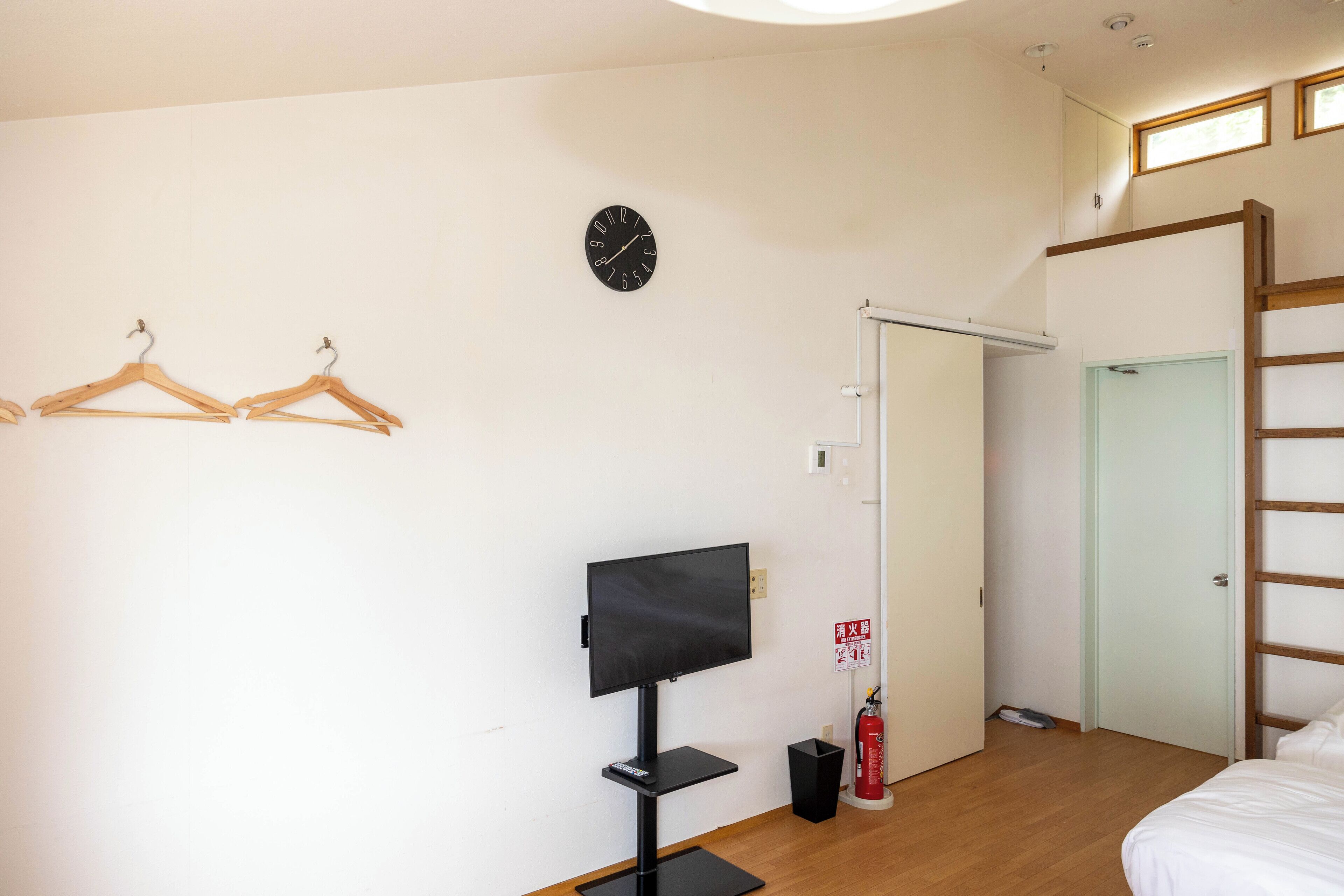 Ett hotellrum med en säng, en tv, trägalgar, en klocka, en brandsläckare och en dörr.