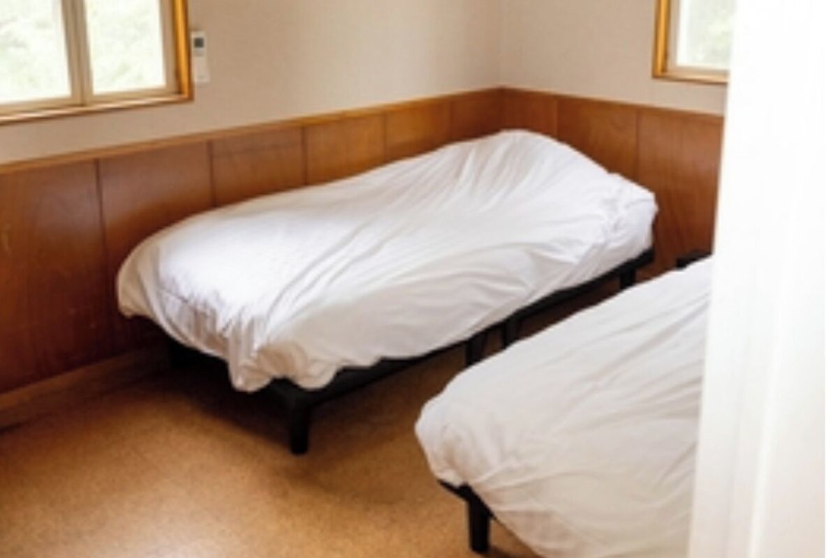 A small, single-room with two single beds, wooden paneling on the walls, and windows providing natural light.