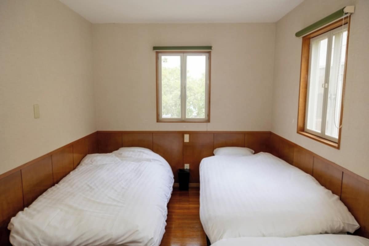 A room with two single beds, wooden headboards, a window with a green frame, and a small trash bin.