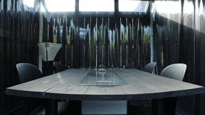 Een ronde tafel met een glazen blad, omringd door vier stoelen, in een kamer met een donkere, spiegelende wand.