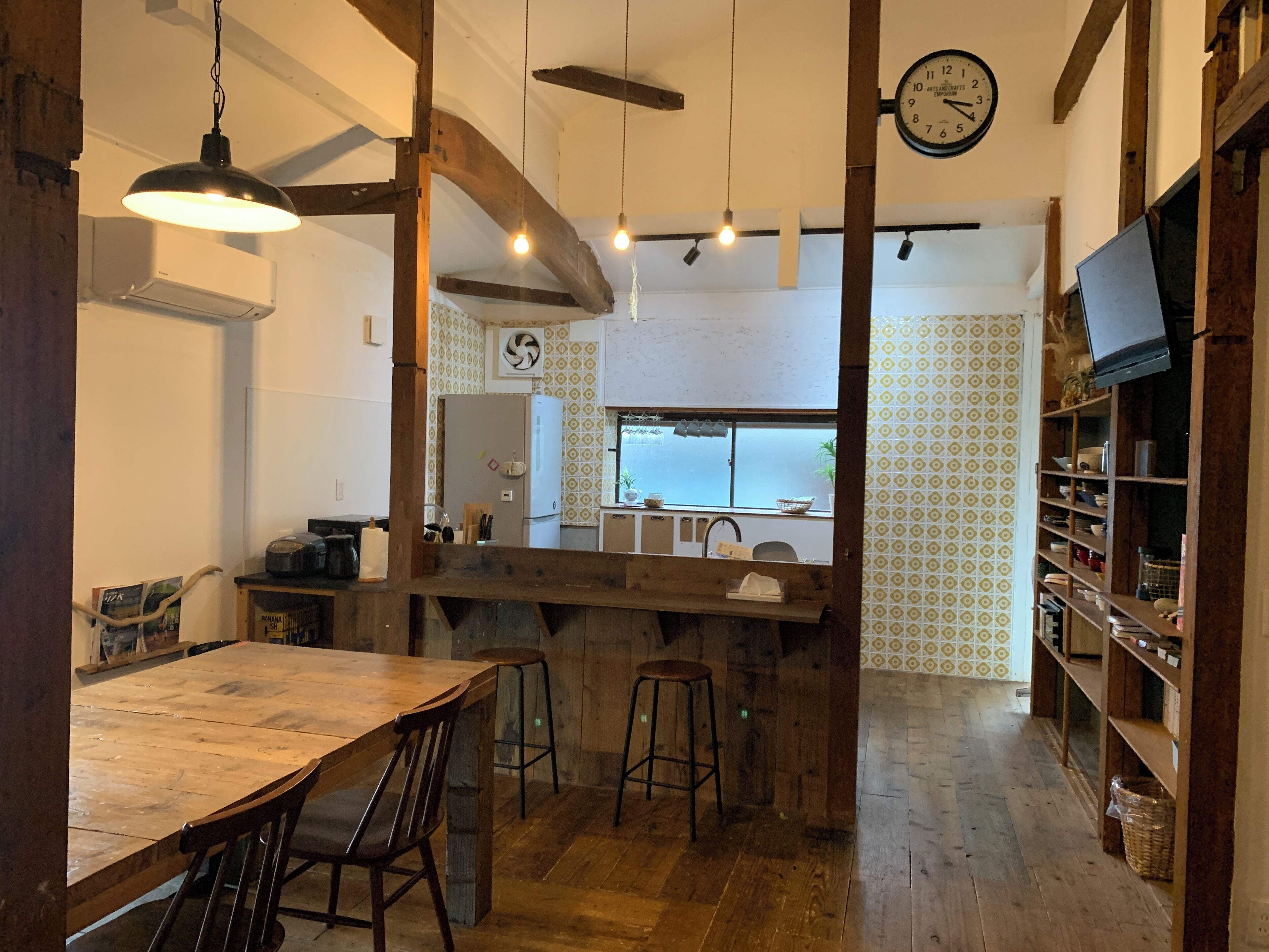A rustic kitchen with wooden beams, a large wooden table, bar stools, and a clock on the wall.