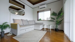 A modern living room with a grey sectional sofa, a round floor lamp, a large mirror, a potted plant, and a window with blinds. - SEA Eirene Hillstop Onna-son (Onna)