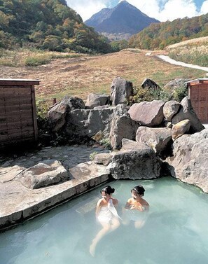 A natural hot spring with two people in the water, surrounded by rocks and a wooden structure. - Pension Yuuzann (Nagano)