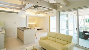 A modern living room with a sofa, a kitchen area with a stove, and a large sliding glass door leading to an outdoor area. - Hummer Village (Kamiamakusa)