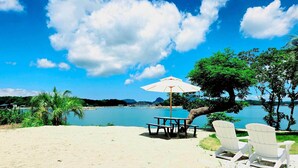 A beach with a table and chairs, a white umbrella, and a view of the water and mountains. - Hummer Village (Kamiamakusa)