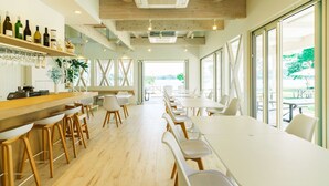 A modern dining area with a bar, white chairs, and large windows offering a view of the outdoors. - Hummer Village (Kamiamakusa)