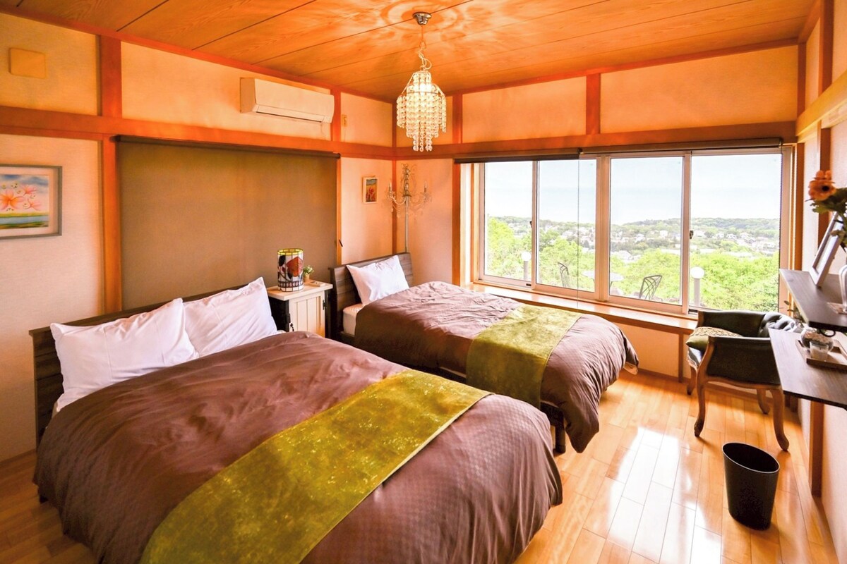 Une chambre avec deux lits, un bureau, une chaise, une poubelle et une grande fenêtre donnant sur un beau paysage.