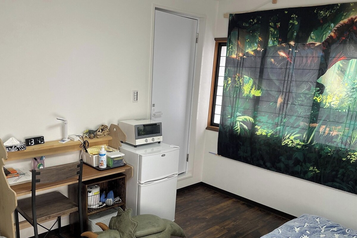 Une petite pièce comprenant un bureau, un réfrigérateur, un four à micro-ondes, une chaise et une grande fenêtre ornée d’un rideau à motifs tropicaux.