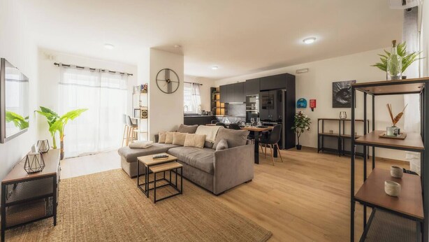 Ein modernes Wohnzimmer mit einer Couch, einem Couchtisch und einem Essbereich mit Stühlen. Im Hintergrund ist eine Küche mit dunklen Schränken und einem Kühlschrank zu sehen.